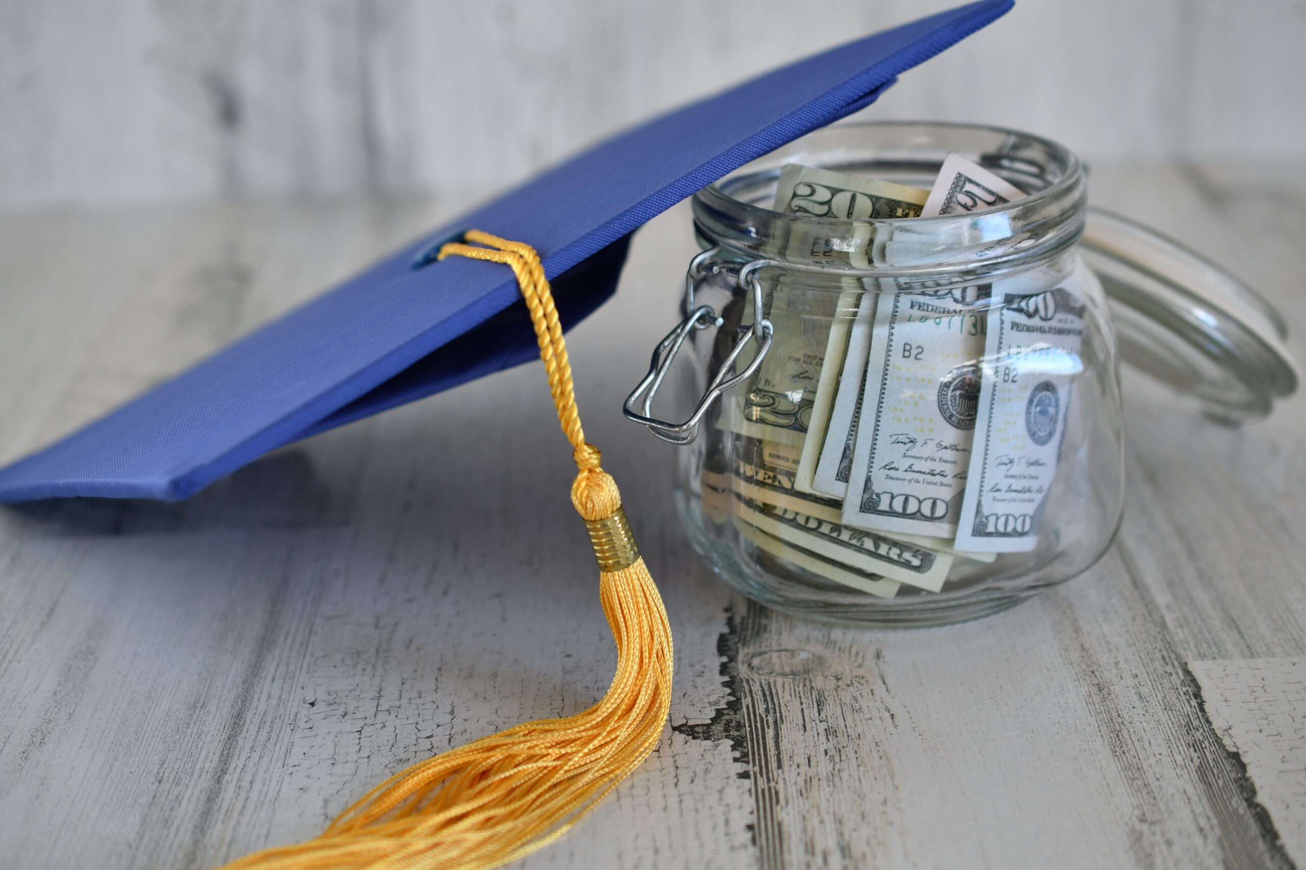 Glass jar filled with cash next to a blue graduation cap, symbolizing financial aid, student benefits, and education funding.